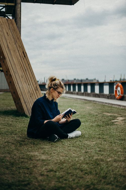 A woman reading a book outdoors beside a quiet stretch of water.